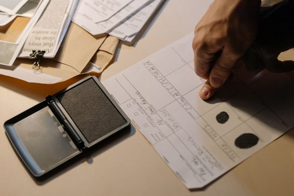 Detalhe de uma mão coletando impressões digitais em um documento sobre uma mesa, com uma almofada de tinta ao lado