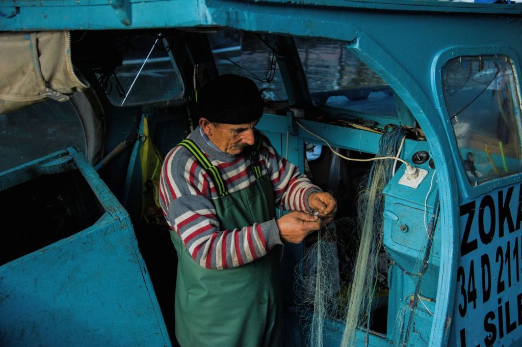 Seleção pública prevê R$ 120 milhões para inovação na pesca artesanal Pescador repara uma rede em um pequeno barco azul.