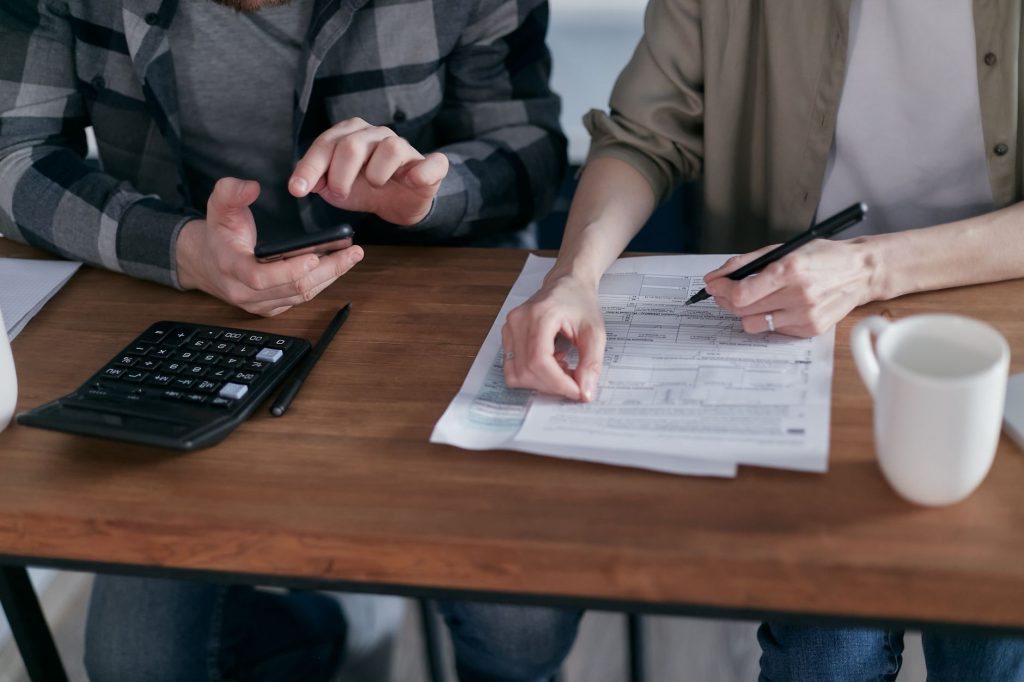 Duas pessoas analisam formulários tributários e fazem cálculos com uma calculadora em uma mesa de trabalho.